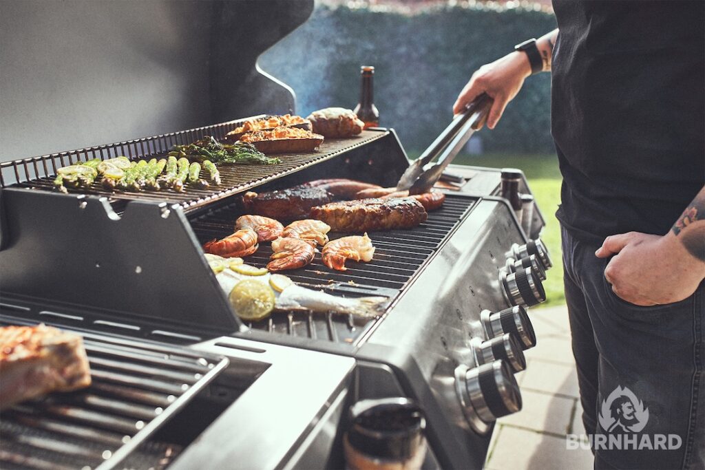 Ein gut gefüllter Gasgrill von der Seite, ein Mann dreht das Fleisch um