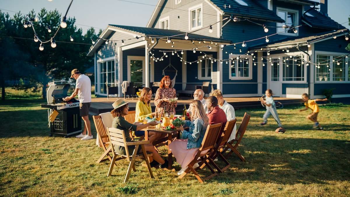 Blick von oben auf eine mehrköpfige Familie an einer langen gedeckten Gartentafel mit Gebäck und Getränken, im Garten hängen Wimpelketten und bunte Luftballons