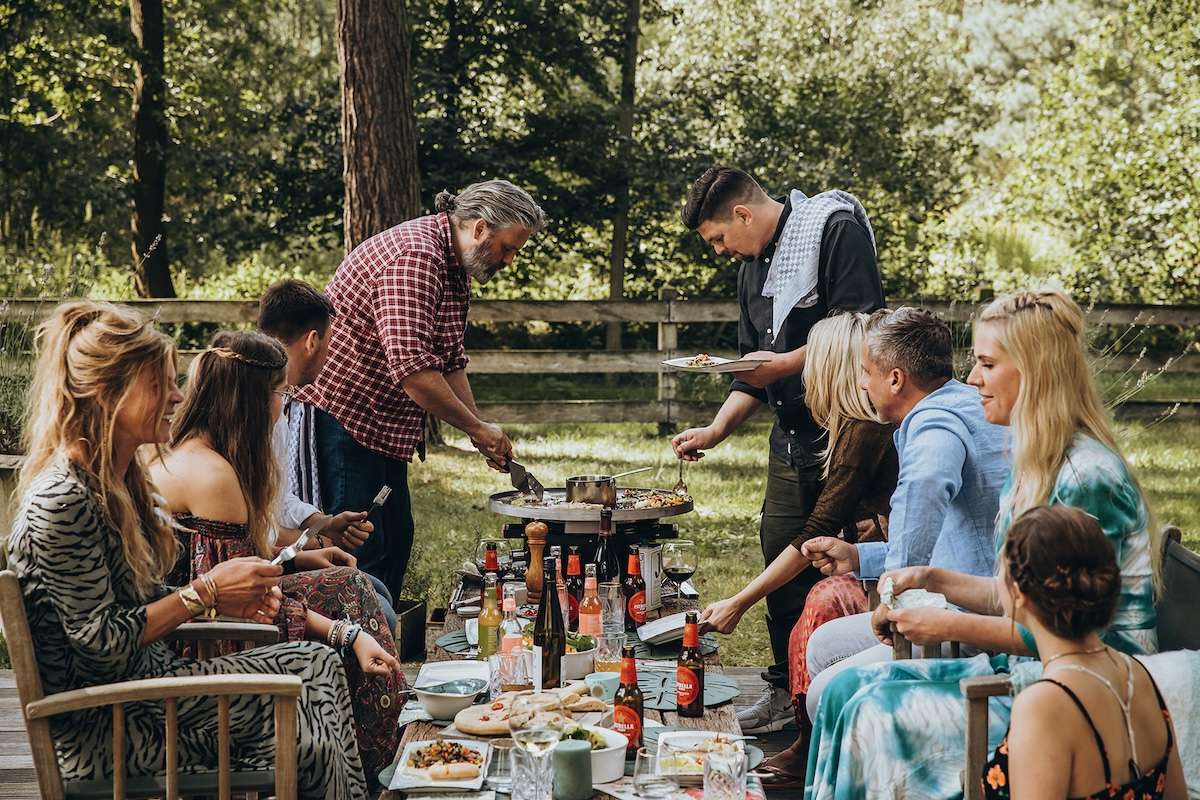 Monolith Feuerplatte im Einsatz auf einer Gartenparty, Freunde an einem langen gedeckten Tisch, zwei Männer bereiten in der Mitte Speisen zu