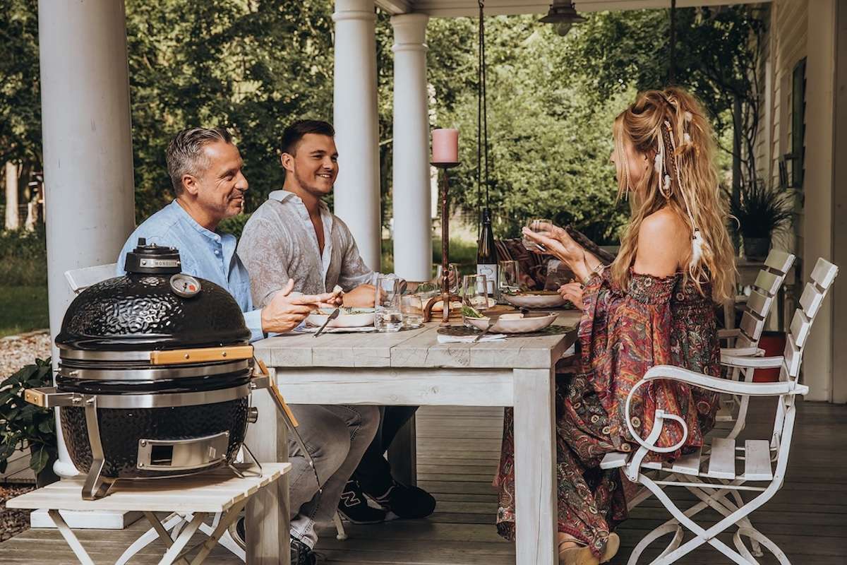 Drei Personen essen gemeinsam an einem Holztisch auf einer überdachten Terrasse, links auf einem Beistelltisch ein kompakter schwarzer Monolith Kamado Grill