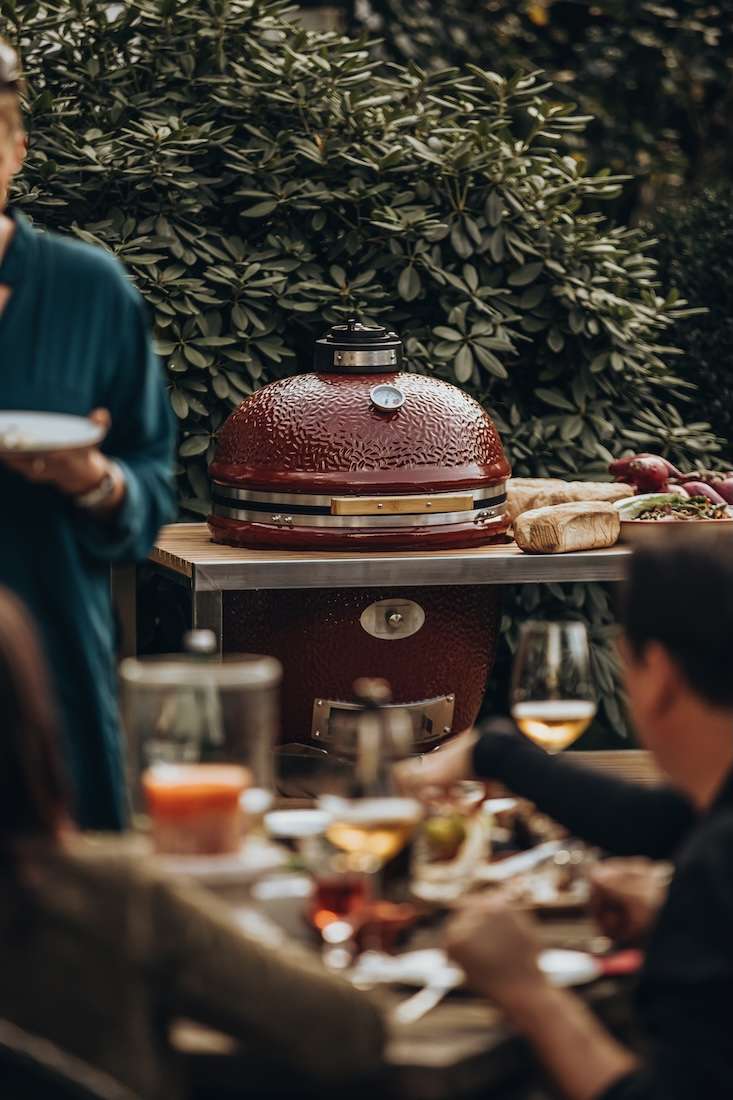 Roter Monolith Kamado Grill aus Keramik auf einem Beistelltisch vor einer Rhododendronhecke, im unscharfen Vordergrund Menschen an einem gedeckten Tisch