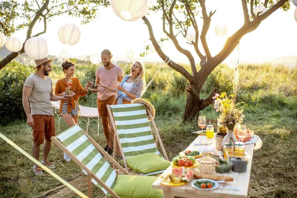 Vier junge Leute stehen bei Sonnenuntergang mit Tellern und Getränken im Garten zusammen, über ihnen Papierlampions, im Vordergrund gestreifte Liegestühle und ein gedeckter Tisch